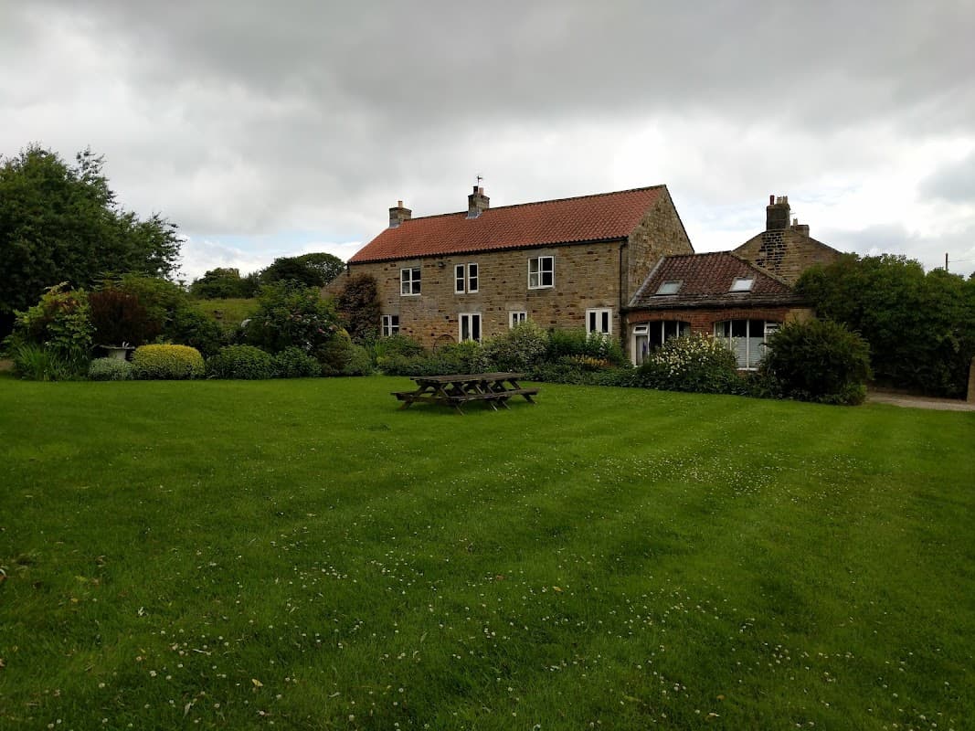 Charming stone hotel with a red-tiled roof, surrounded by lush green gardens and picnic tables under a cloudy sky.