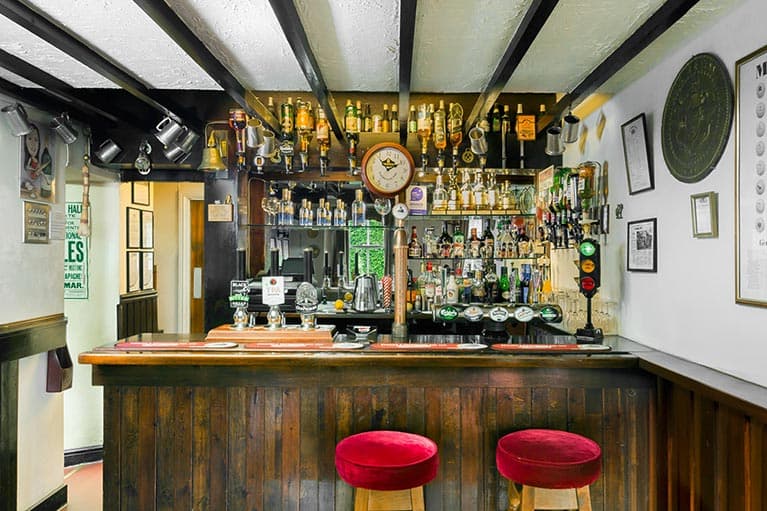 Wooden bar with red stools, shelves lined with bottles, and a clock on the wall in a cozy pub setting.