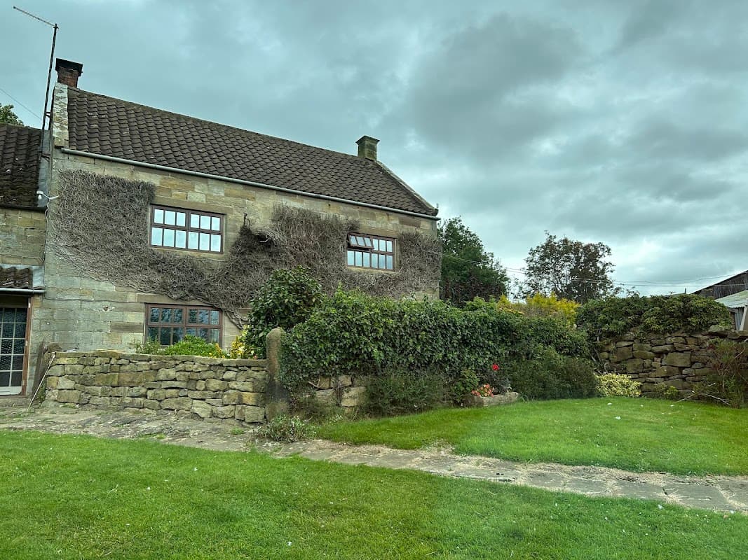 Stone cottage with ivy, surrounded by green grass and a stone wall, under a cloudy sky in Lealholm, Yorkshire.