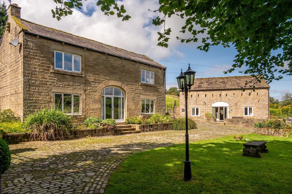 Two stone cottages with large windows, surrounded by greenery and a cobblestone path, in a scenic Yorkshire setting.