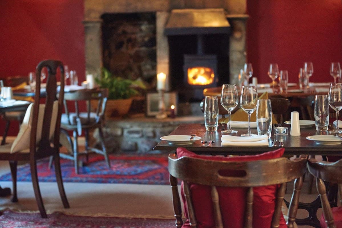 Cozy dining area with wooden tables, elegant glassware, and a warm fireplace against a rich red wall.