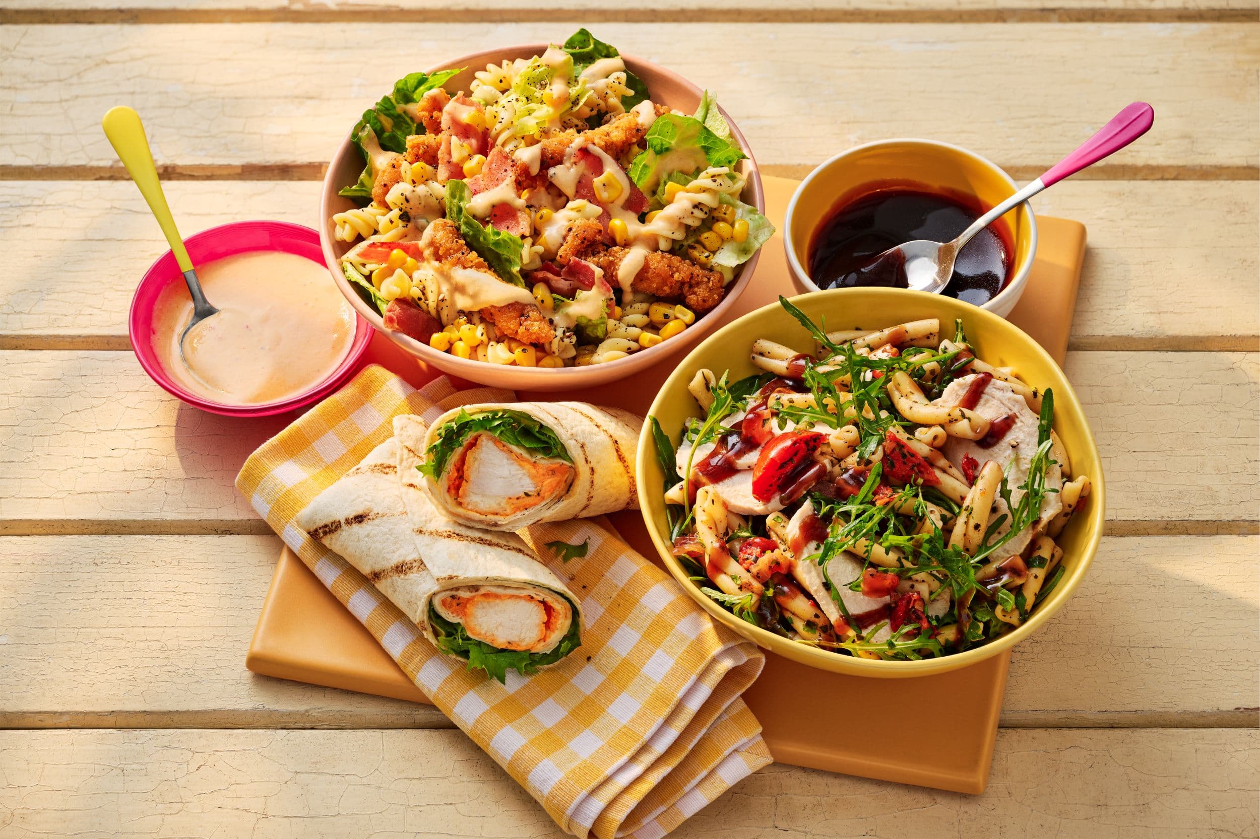 Two colorful salads with sauces and a chicken wrap on a table