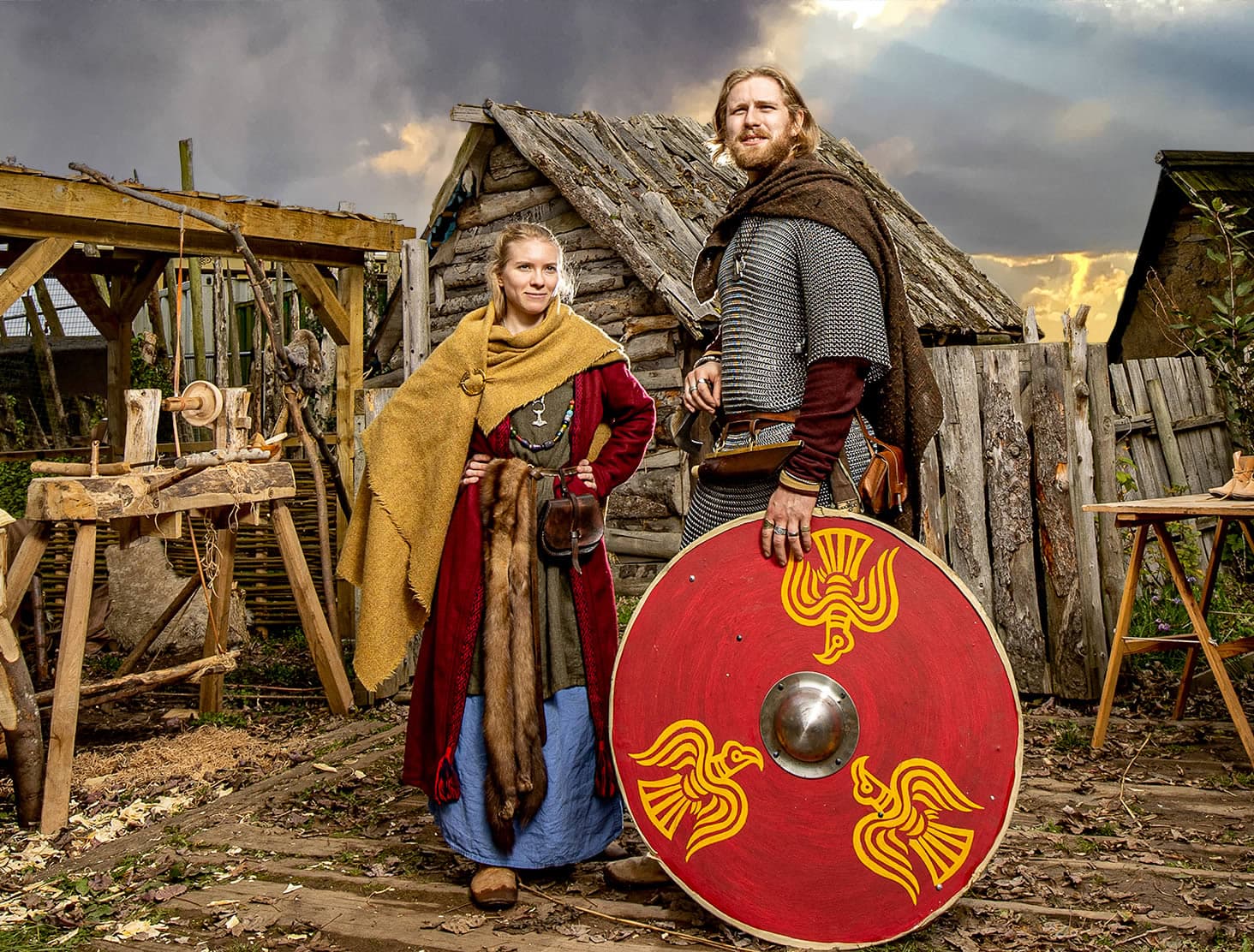 Two people in Viking attire, standing on wooden planks, with a rustic wooden hut and tools in the background.