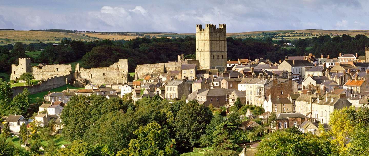 Richmond Castle rises above a river, framed by lush greenery and a stone bridge, under a blue sky with scattered clouds.
