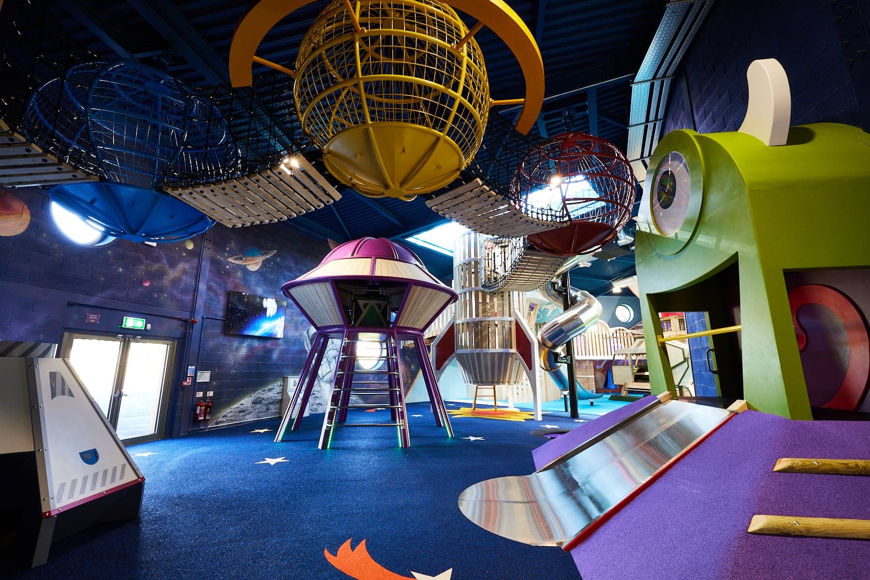 Colorful indoor playground with space-themed structures, slides, and climbing nets under a high blue ceiling.