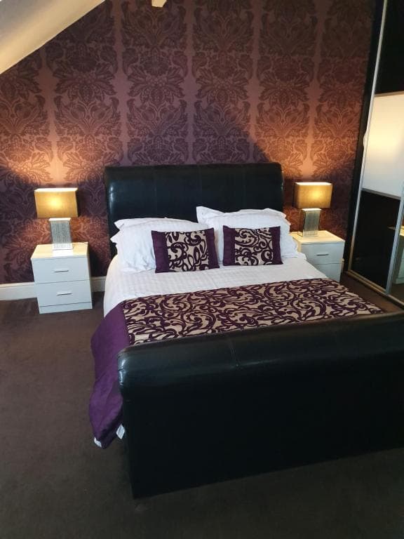 Cozy bedroom with a dark leather bed, purple damask wallpaper, matching bedding, and two bedside lamps on white tables.