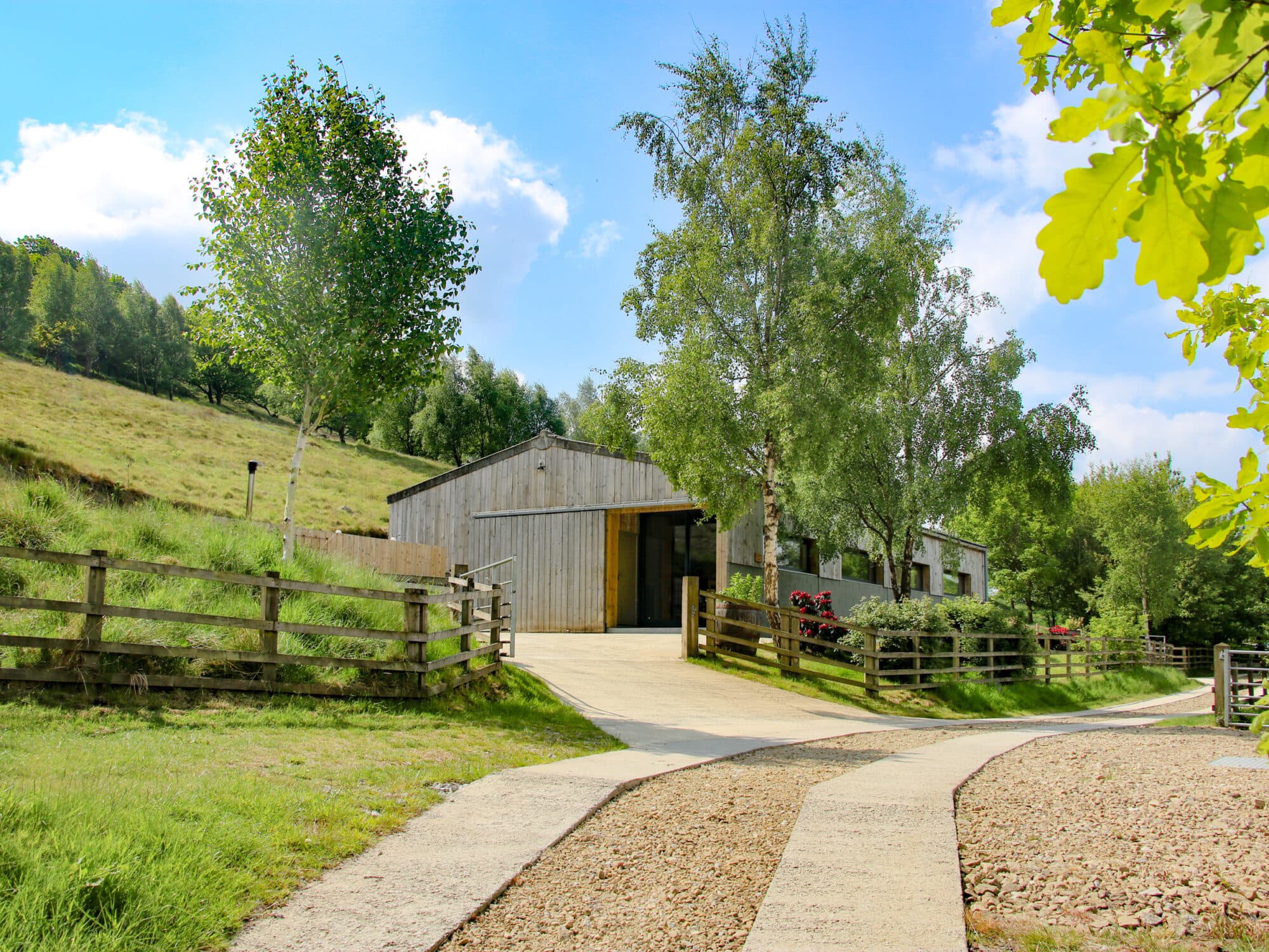 Rustic wooden barn surrounded by lush trees and rolling grassy hills, with a gravel path leading to the entrance.