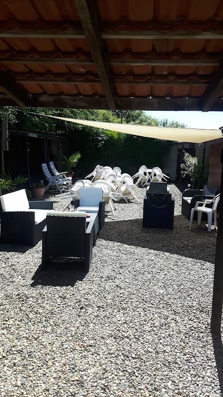 Outdoor seating area with lounge chairs and tables on gravel, surrounded by greenery and shaded by a canopy.