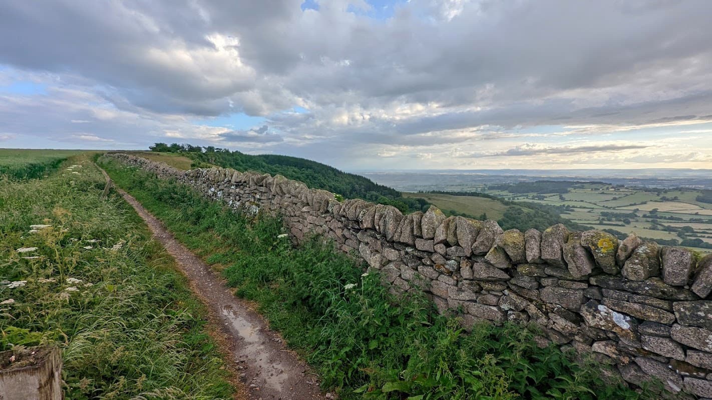 A gravel path alongside a dry stone wall, with green fields and rolling hills under a cloudy sky in Boltby, Yorkshire.
