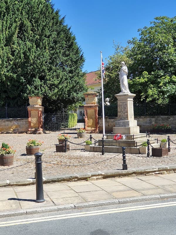 Free parking area with a statue, flower pots, and trees under a clear blue sky in Boroughbridge, Yorkshire.