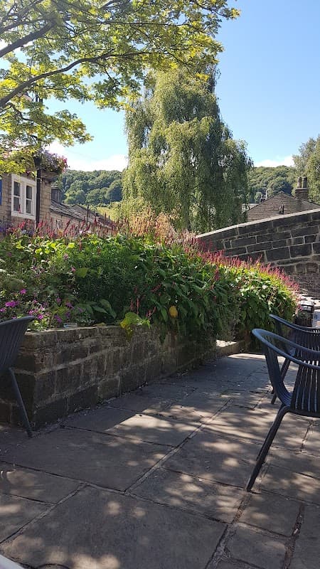 Lush greenery and colorful flowers surround a stone wall, with blue sky and trees in the background. Outdoor seating visible.