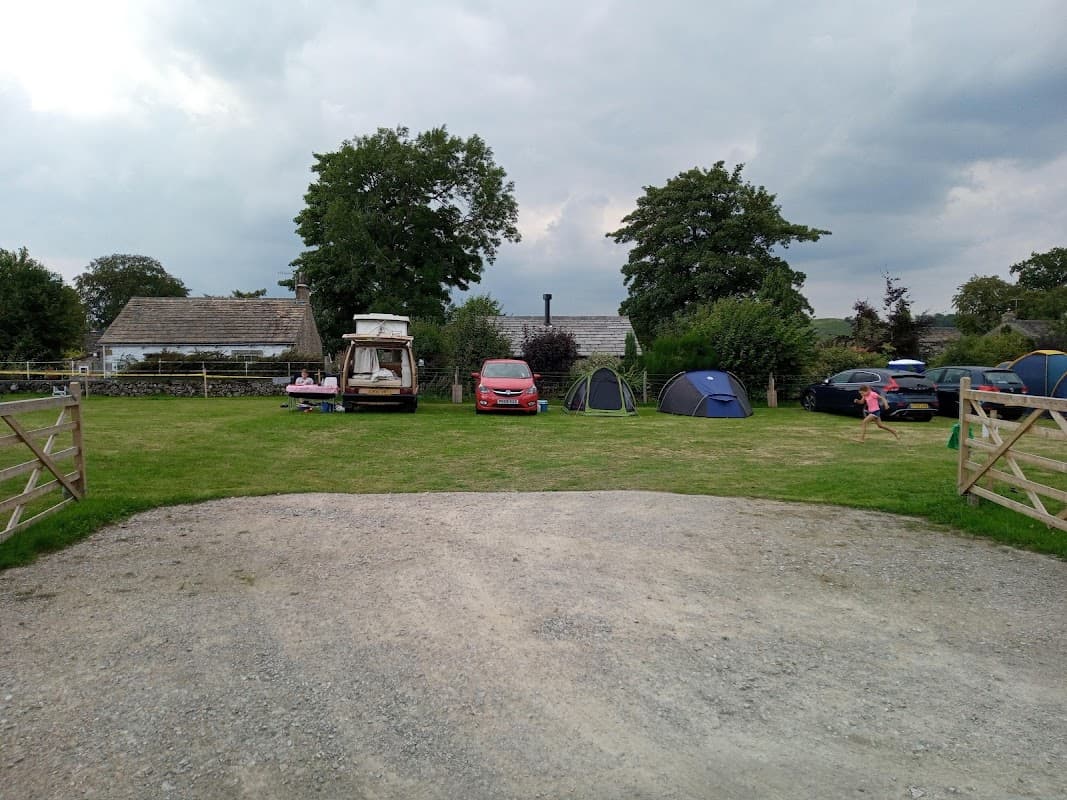 A grassy area with tents, parked cars, and a caravan, surrounded by trees and a stone building under a cloudy sky.