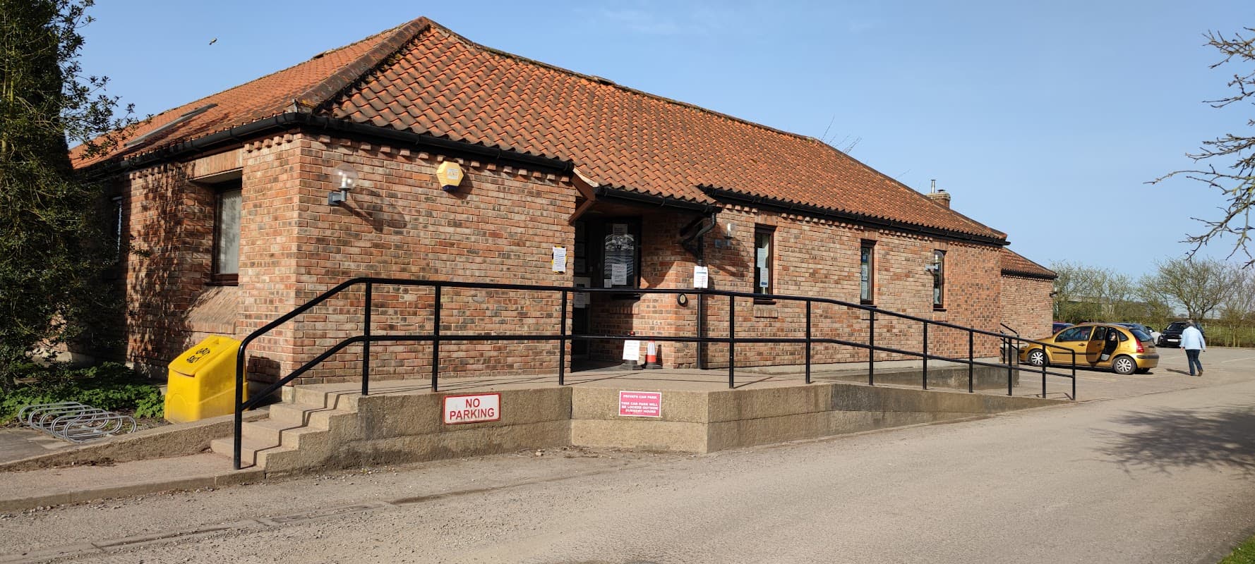 Brick building with a red tiled roof, ramp entrance, and a sign indicating no parking. A taxi is parked nearby.