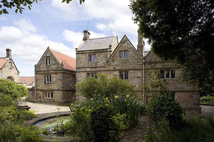 Historic stone building surrounded by lush greenery and a garden, with a blue sky and scattered clouds above.