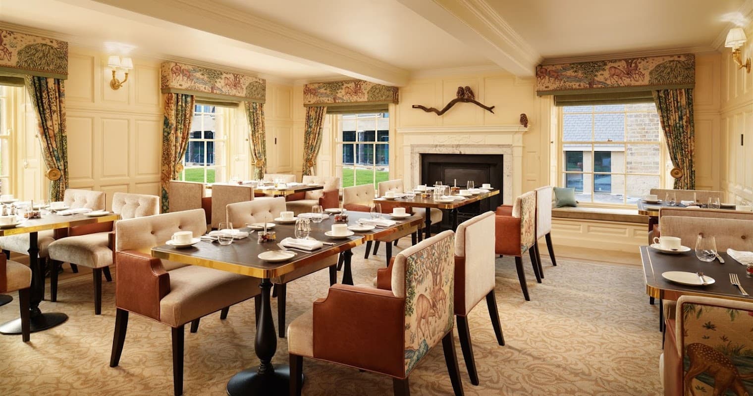 Elegant cafe interior with wooden tables, plush chairs, and large windows, featuring a cozy fireplace and tasteful decor.