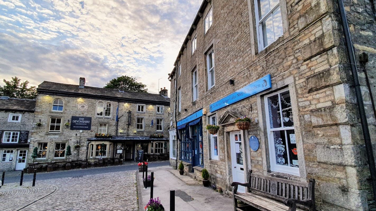 Quaint stone buildings surround Grassington Square, featuring shops, benches, and a charming sky overhead.