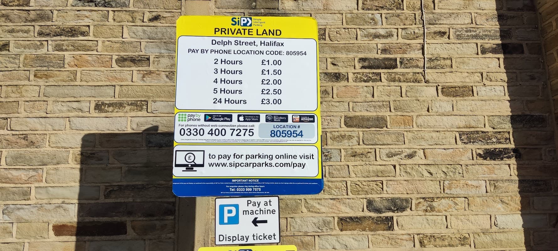 Sign detailing parking rates and payment options at Delph Street car park, with a stone wall background.