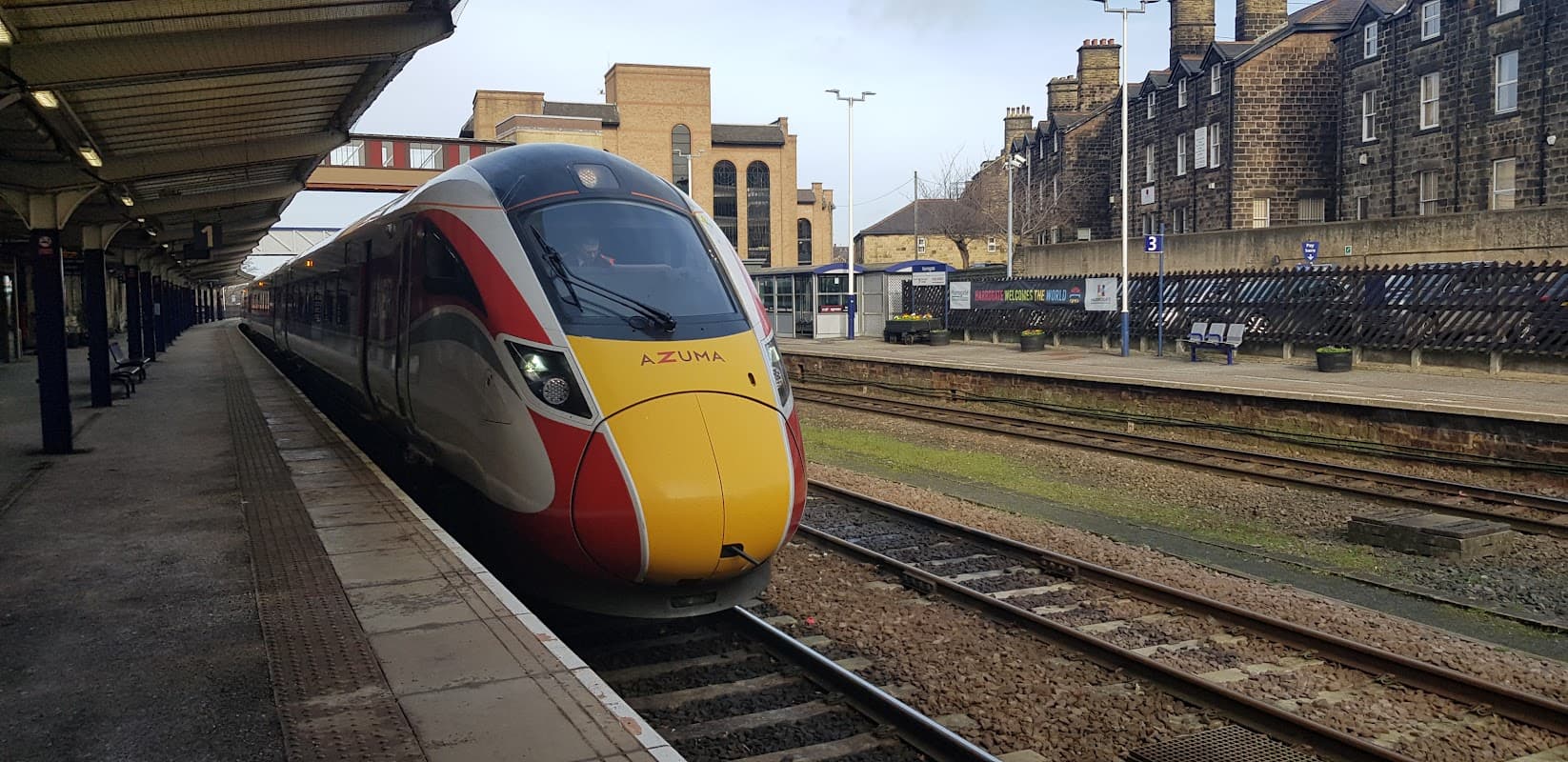 A red and yellow train at Harrogate railway station, with stone buildings and tracks visible in the background.