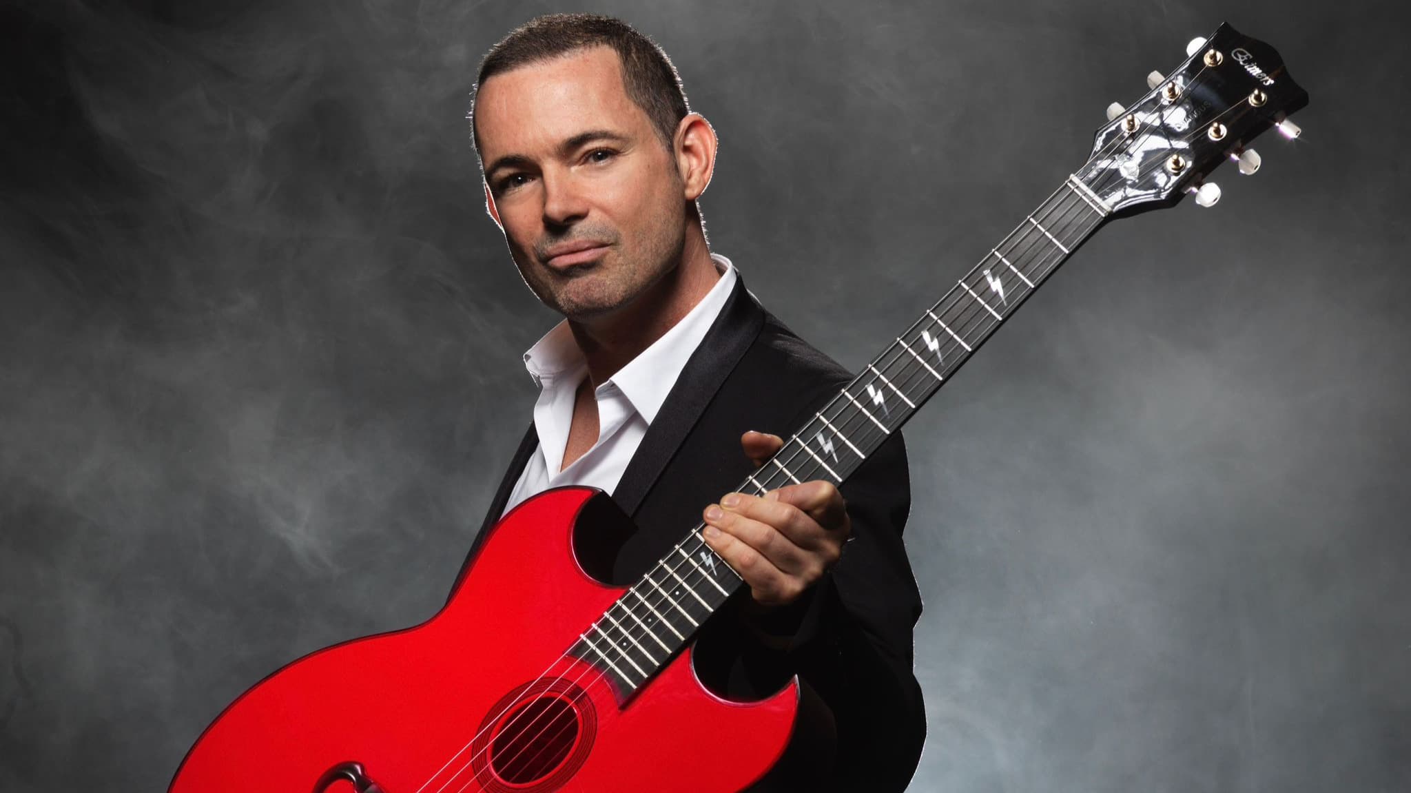 Man holding a red electric guitar against a dark, smoky background
