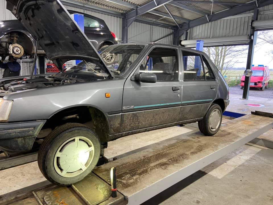 A grey car on a lift in a garage, with tools and other vehicles visible in the background.
