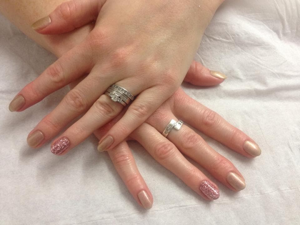 Two hands with manicured nails, one featuring glitter, adorned with multiple rings, resting on a white surface.