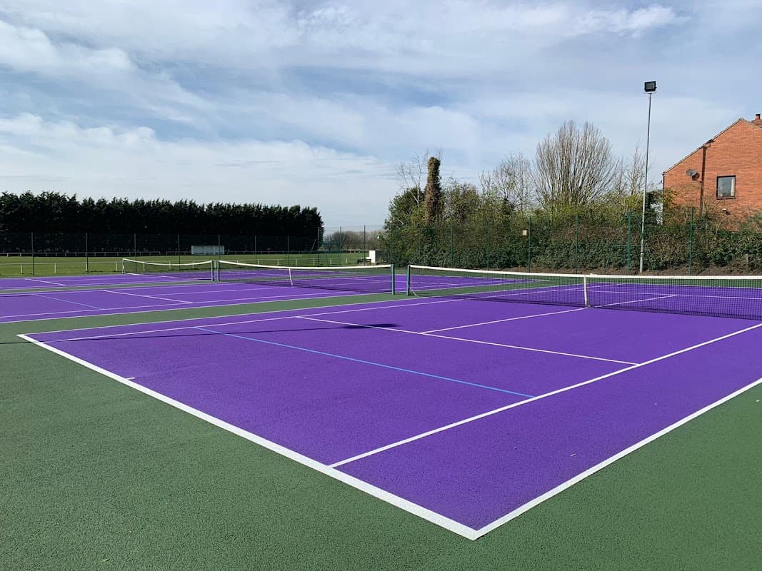 Purple tennis courts with white lines, surrounded by greenery and a brick building in the background.
