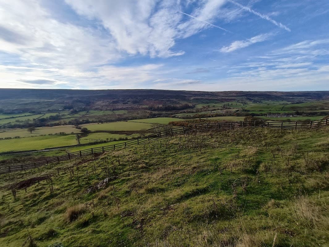 Rolling green hills under a blue sky with wispy clouds, bordered by a wooden fence in Rosedale Abbey, Yorkshire.