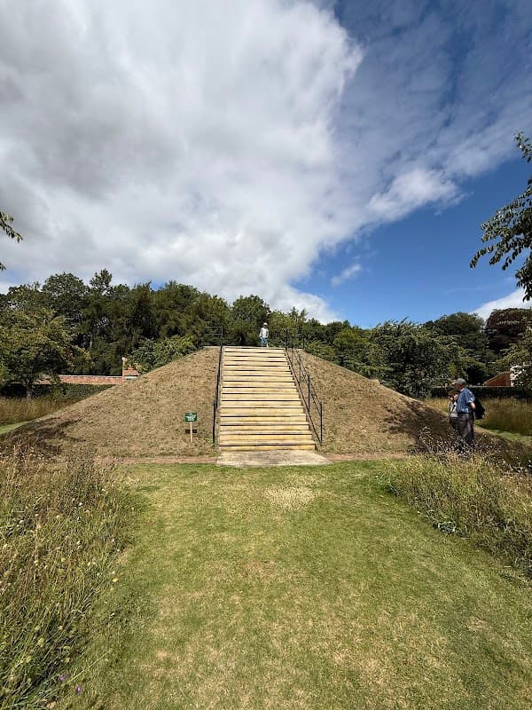 Staircase leading up a grassy mound, surrounded by lush greenery and under a partly cloudy sky.