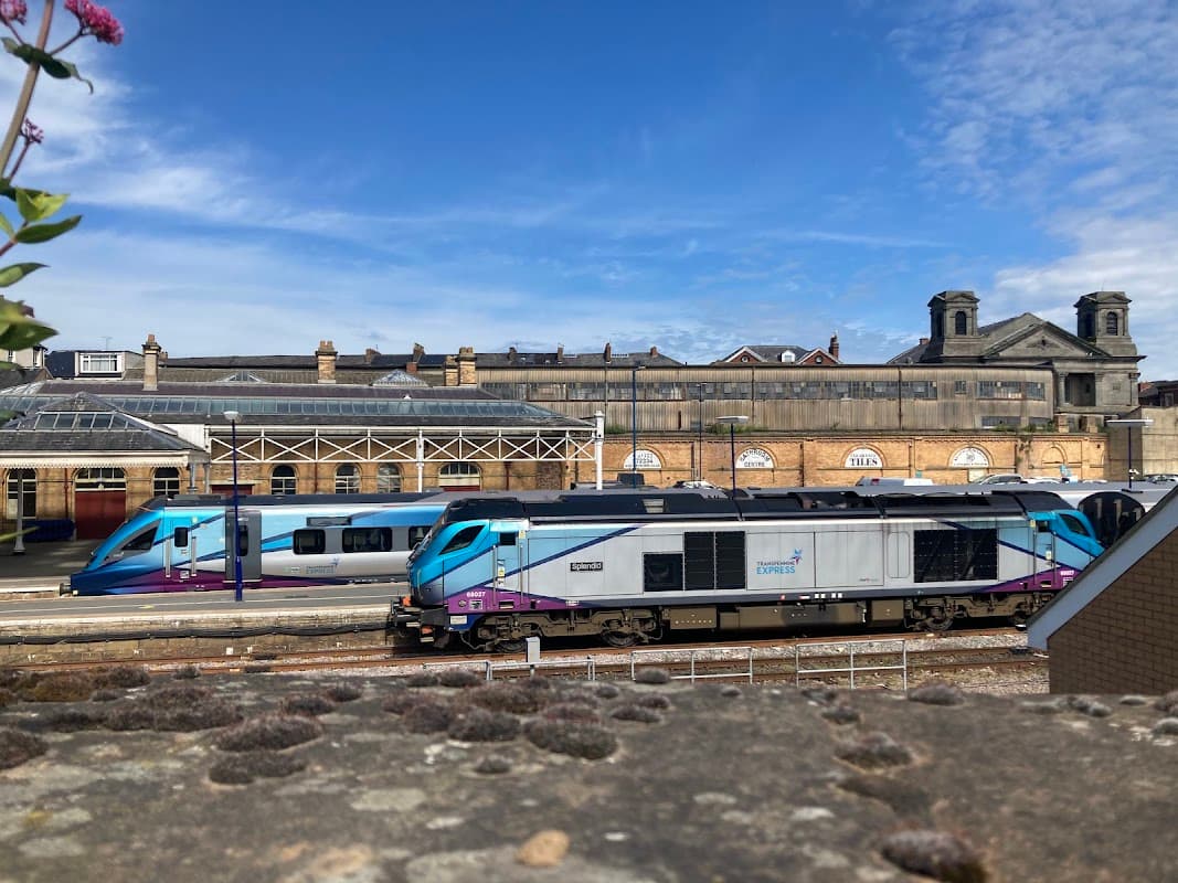 Two trains are stationed at Scarborough Railway Station, with a historic building and blue sky in the background.