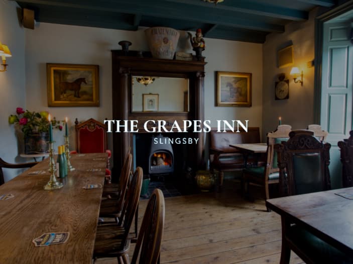Cozy interior of The Grapes Inn with wooden tables, chairs, a fireplace, and framed artwork on the walls.