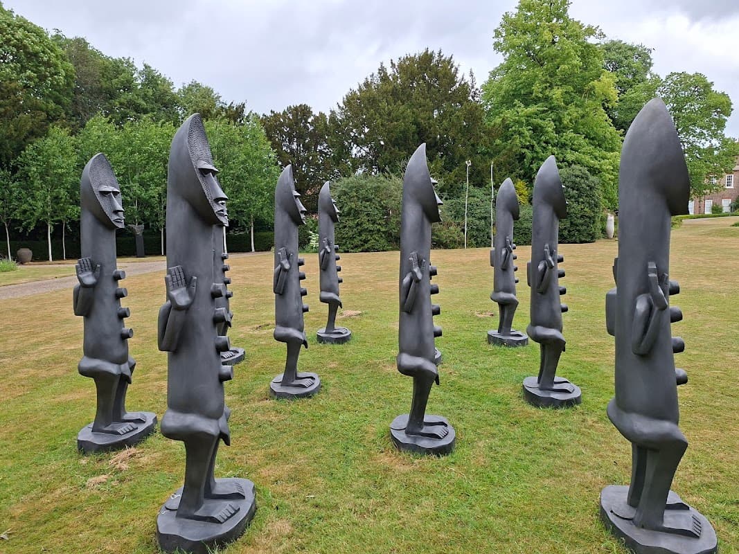 A circular arrangement of tall, abstract black sculptures on green grass, surrounded by trees and a cloudy sky.