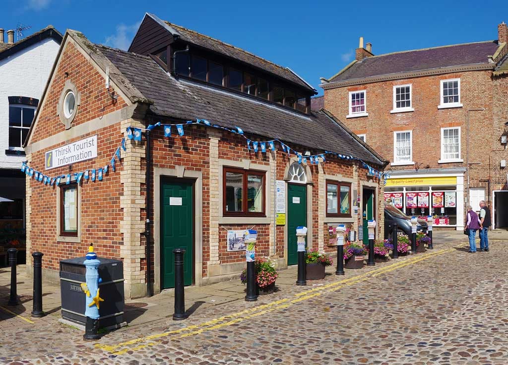 Brick building with green doors, bunting, and flower pots, located in a cobbled street in Thirsk, Yorkshire.