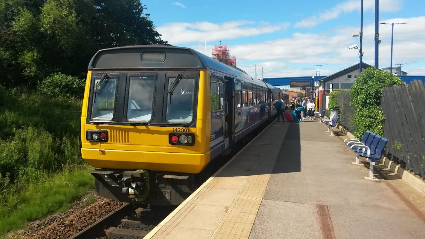 A yellow and blue train at Thornaby station, with passengers waiting on the platform and greenery in the background.