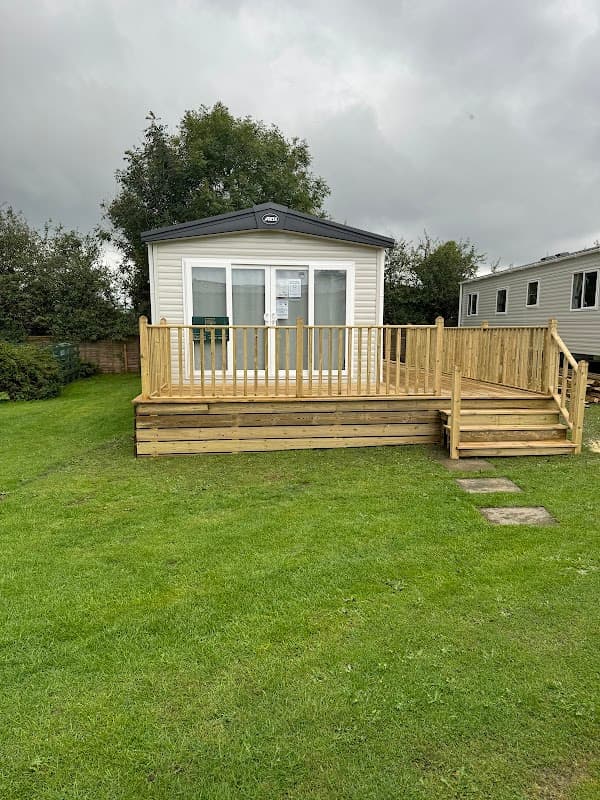 Modern caravan with a wooden deck, surrounded by grassy areas and trees under a cloudy sky.