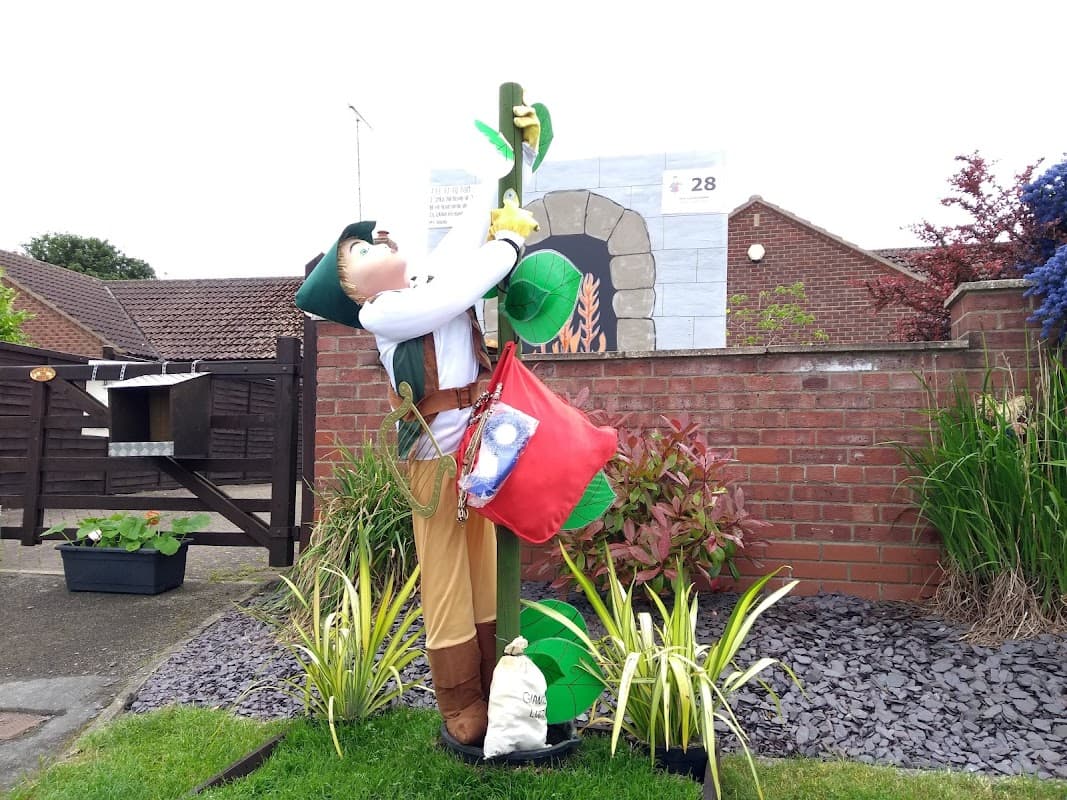 A scarecrow figure climbing a post, wearing a green hat, with a red bag and surrounded by plants in Wetwang.