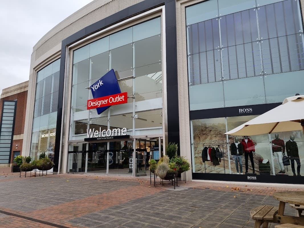 Entrance to Designer Outlet York with large glass windows, "Welcome" sign, and displayed clothing on mannequins.