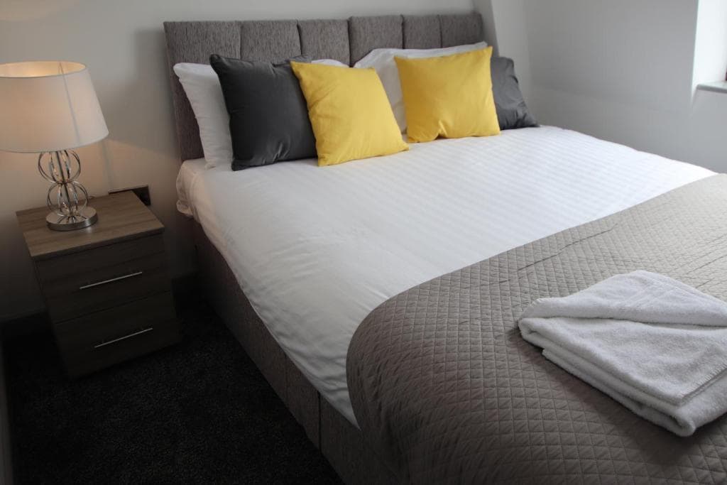 Modern bedroom with a grey headboard, yellow cushions, bedside lamp, and folded towels, Halifax, Yorkshire.