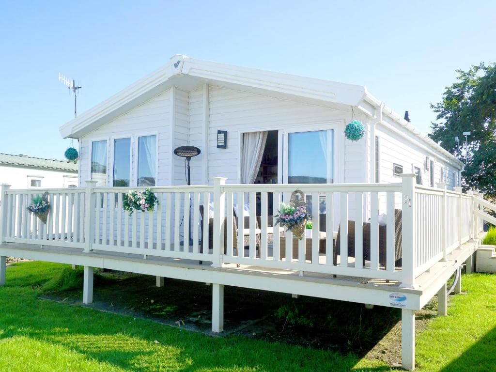 White cottage with large deck, surrounded by green lawn in sunny Cayton, Yorkshire. Hanging flower baskets add charm.
