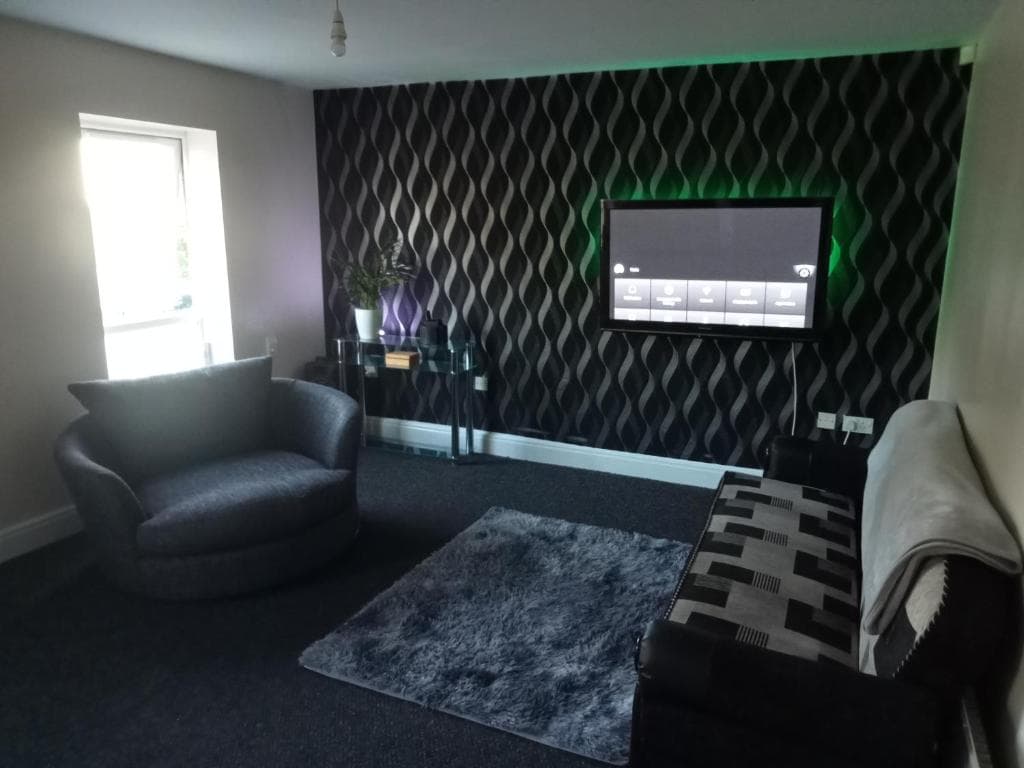 Spacious living room in Dudley Hill apartment, featuring modern decor, patterned wallpaper, plush seating, and a wall-mounted TV.