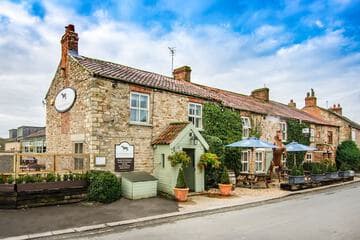Charming stone inn with ivy-covered walls, chimney stacks, and outdoor seating under blue umbrellas on a country road.
