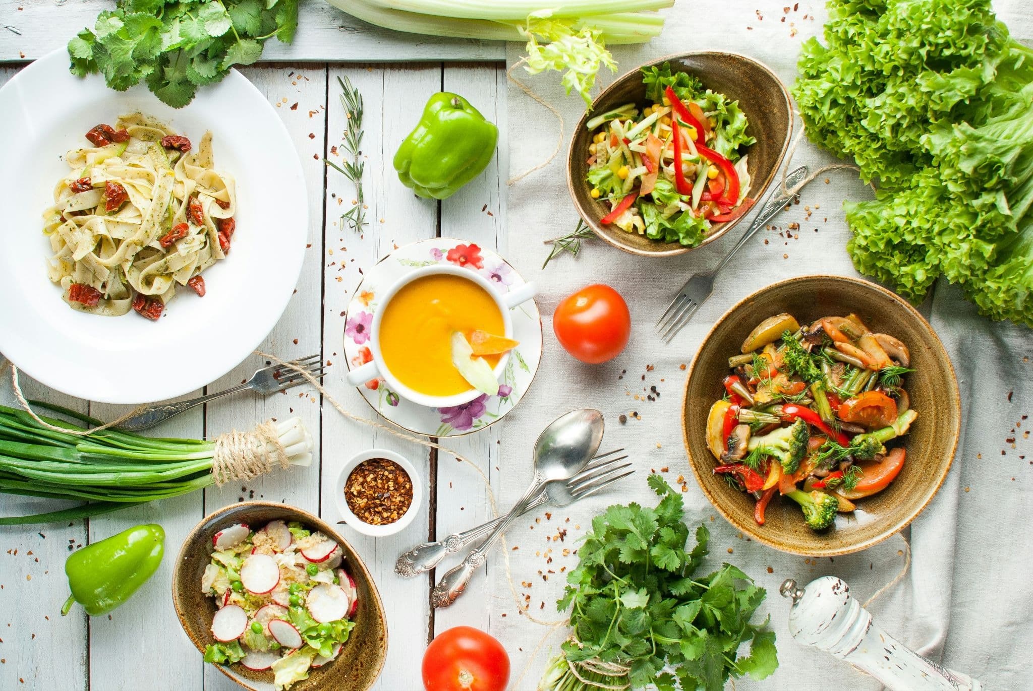 Vibrant spread of plant-based dishes, including pasta, salads, and soup, surrounded by fresh vegetables on a rustic table.