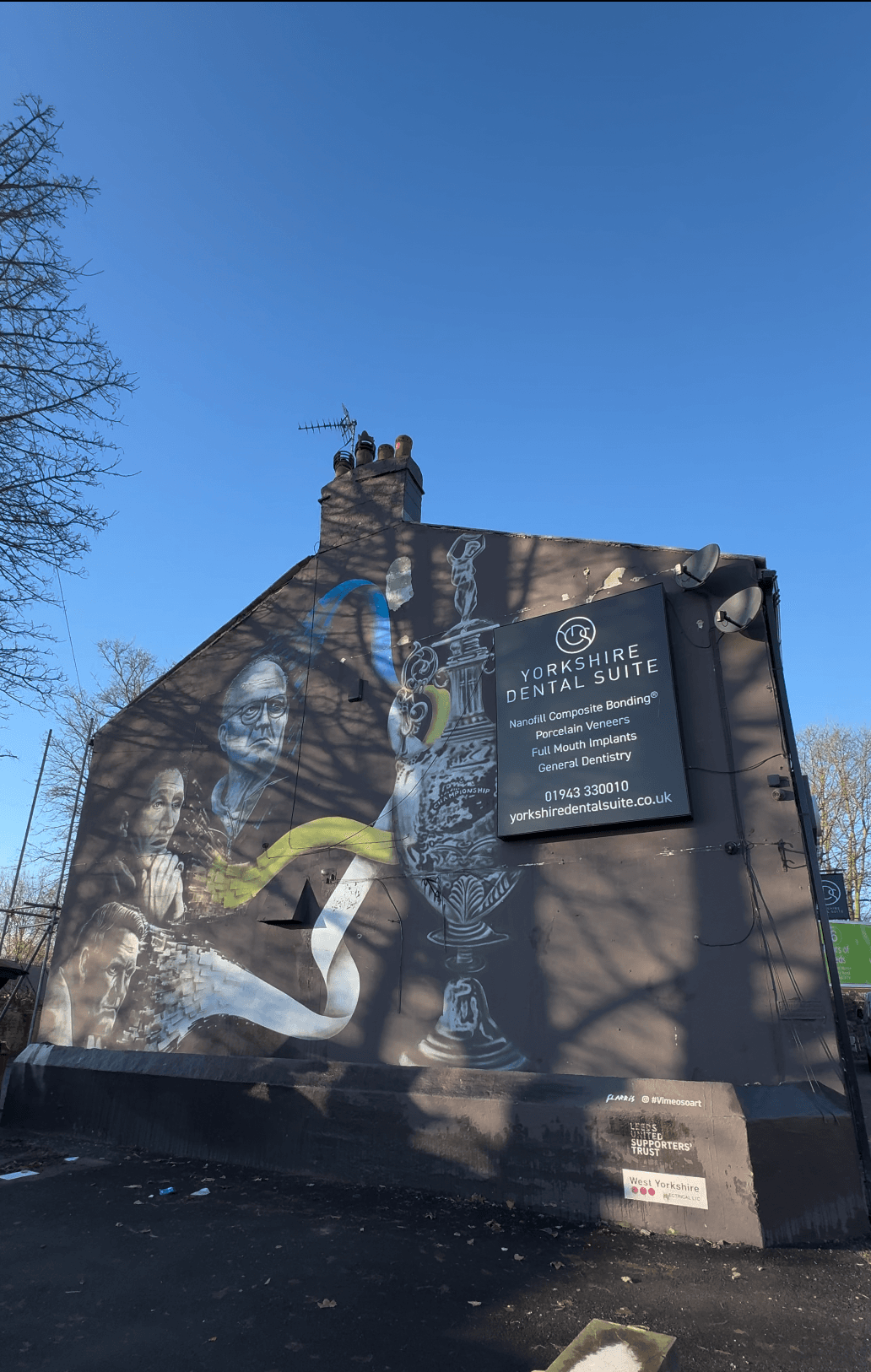 A vibrant mural depicting Leeds United themes on the side of Yorkshire Dental Suite in Guiseley, with workers on the roof.