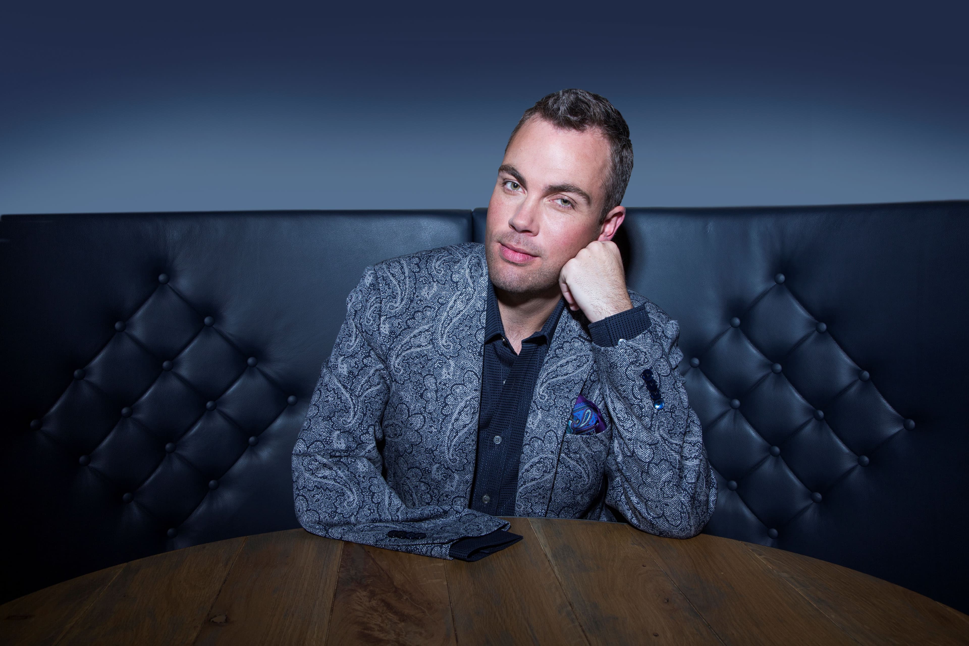 A stylish concert pianist poses thoughtfully at a table, showcasing a modern interior with plush seating and warm lighting.