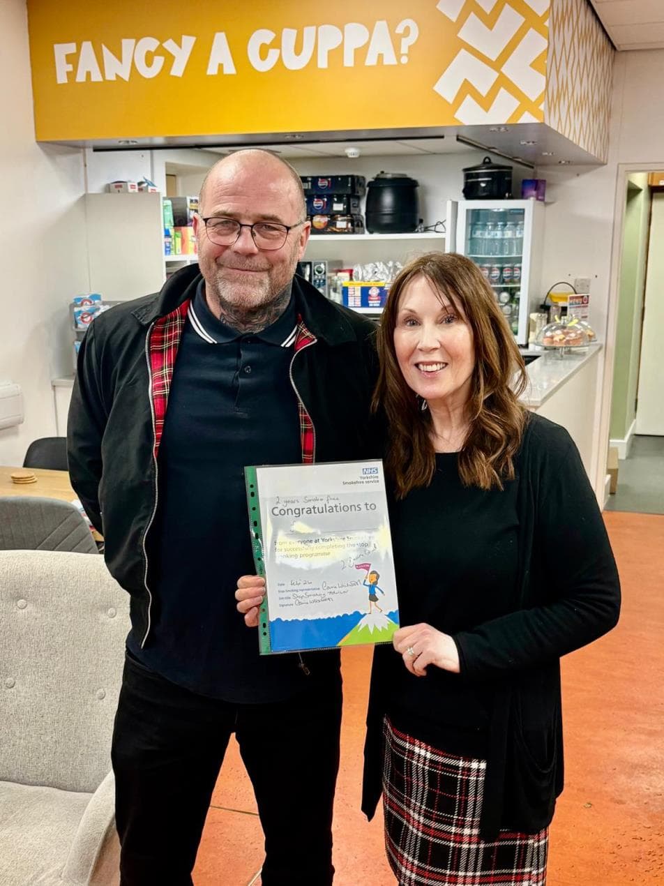 Two smiling individuals hold a certificate in a cozy community space with a kitchen area and a "Fancy a cuppa?" sign.