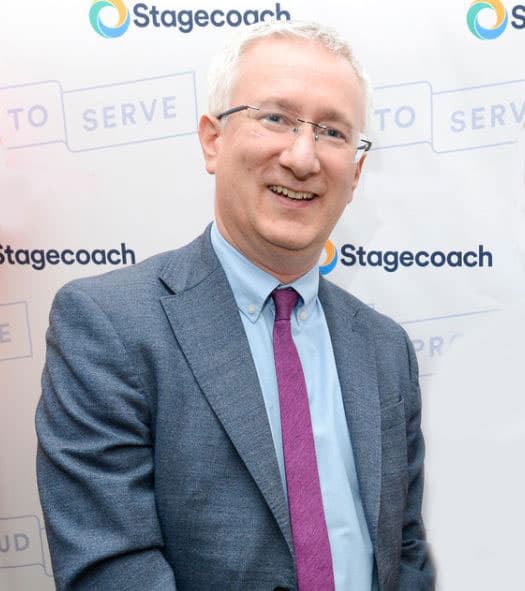 Smiling man in a suit stands against a Stagecoach backdrop, promoting community engagement and fair work initiatives.