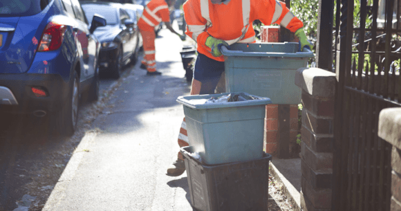 Two workers in high-visibility clothing collect wheeled recycling bins from a residential street in Yorkshire.