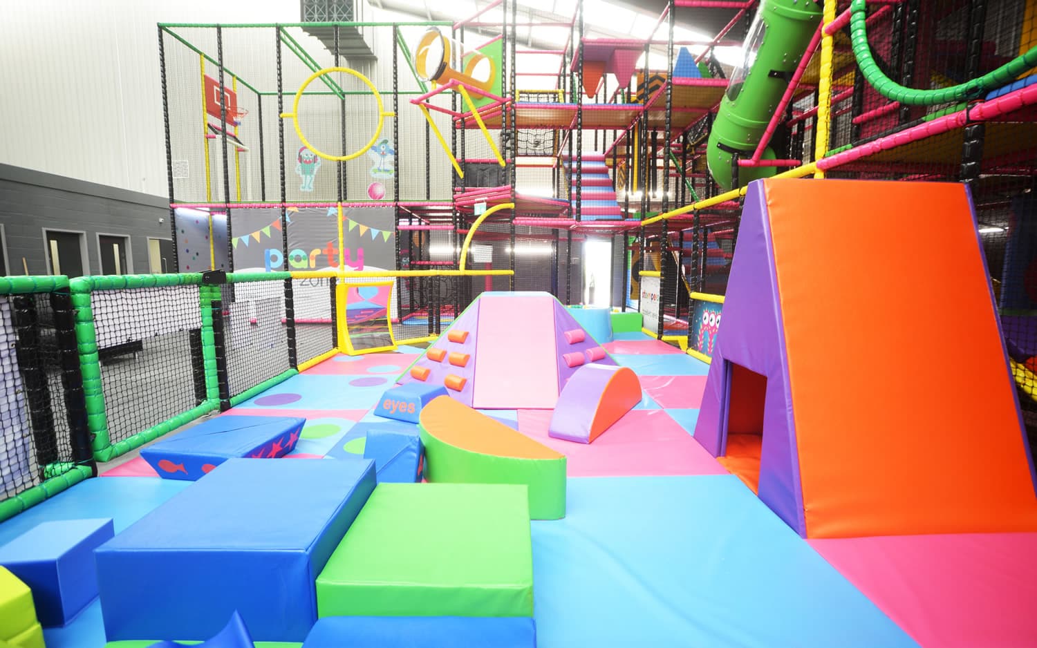Vibrant indoor play area with colorful climbing structures, slides, and padded obstacles under a high ceiling.