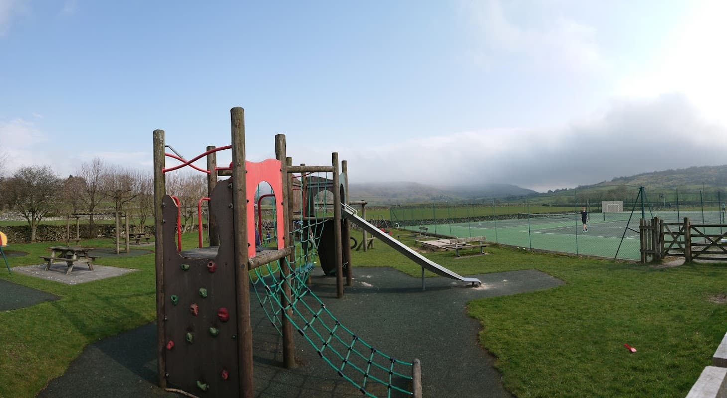 Colorful play equipment with a climbing frame and slide, surrounded by green grass and distant hills under a cloudy sky.