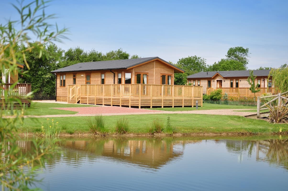 Wooden lodges with decks by a tranquil pond, surrounded by greenery in Balk, North Yorkshire.