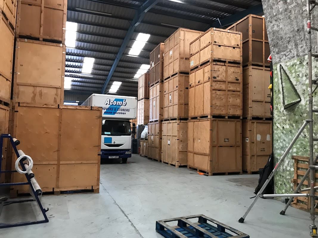 Wooden storage crates stacked in a warehouse with a removal truck labeled "Acomb" in the background.